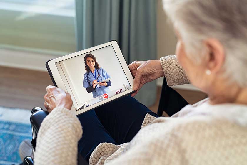 Back view of senior woman sitting in wheelchair making video call with her doctor while staying at home during covid pandemic. Close up of helpful general practitioner in video conference with old woman on digital tablet. Sick woman in online consult