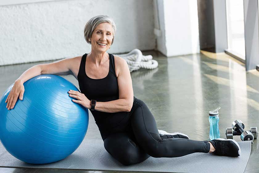 Full length of happy senior woman in black leggings and tank top sitting near fitness ball in gym