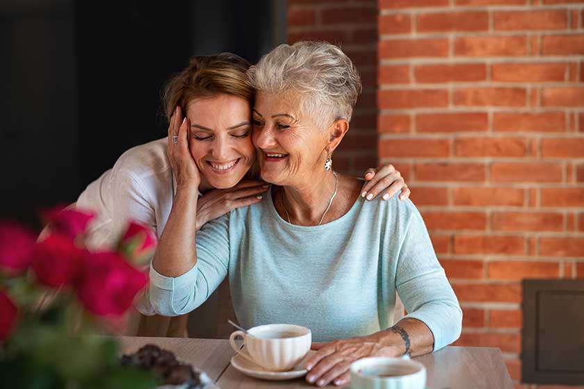Happy senior mother embracing with adult daughter when sitting and having coffee indoors at home.